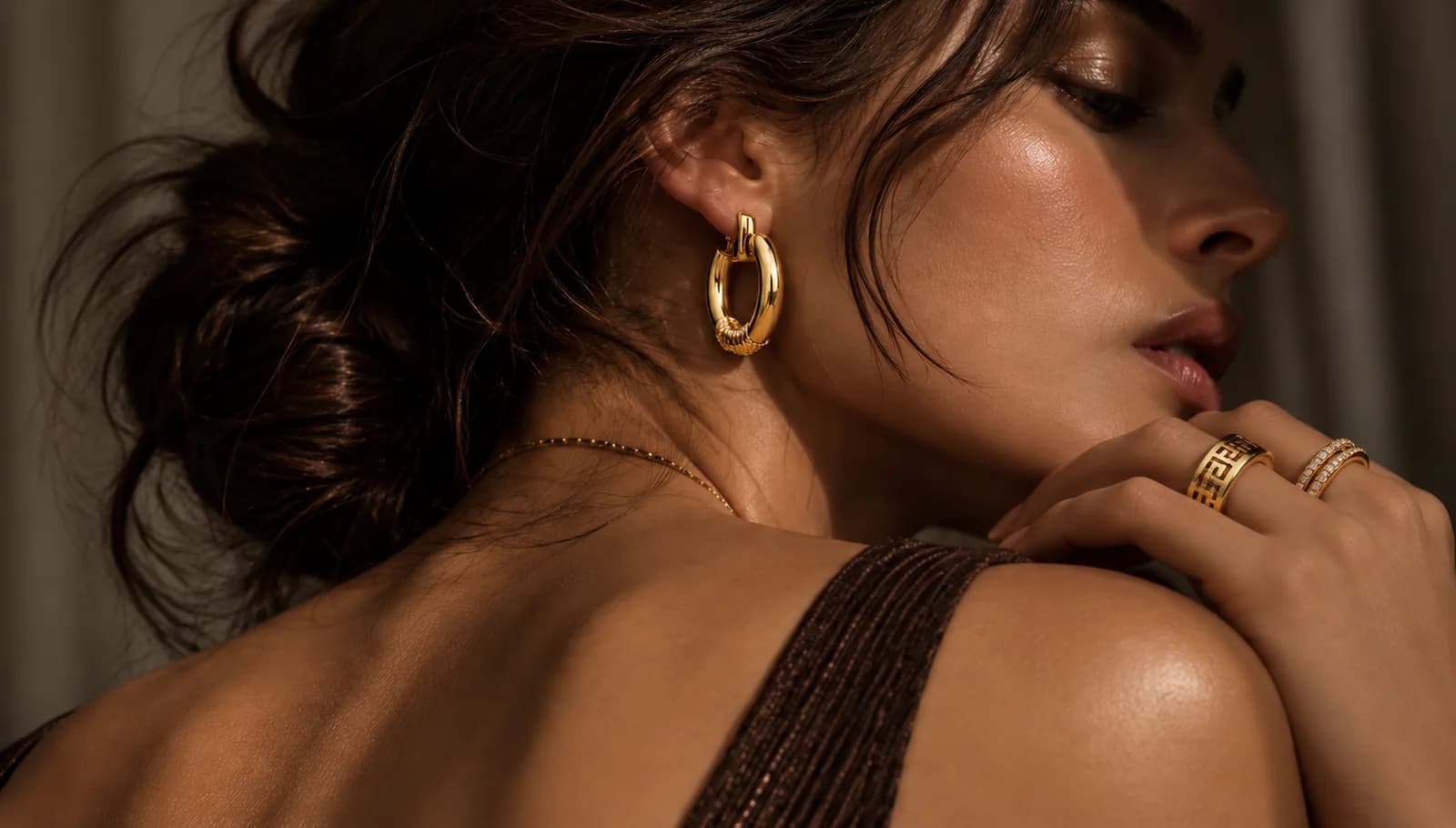 A woman with her hair swept up, wearing a wide gold hoop earring, a fine gold chain and two gold rings, photographed in warm low light